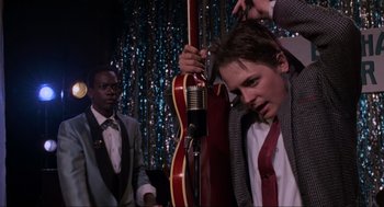 Movie still from “Back to the Future Part II” (1989), directed by Robert Zemeckis – A young man is holding a guitar and a microphone; Medium shot, Over the shoulder angle