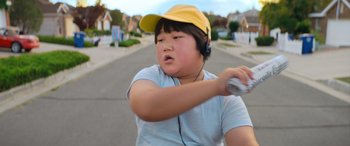 Movie still from “Barb and Star Go to Vista Del Mar” (2021), directed by Josh Greenbaum – A young boy wearing headphones and a yellow hat; Close Up shot, Low angle