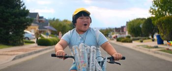 Movie still from “Barb and Star Go to Vista Del Mar” (2021), directed by Josh Greenbaum – A young boy riding a bike with a basket full of water bottles; Medium shot, Low angle