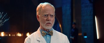 Movie still from “Barb and Star Go to Vista Del Mar” (2021), directed by Josh Greenbaum – An older man in a white lab coat and bow tie; Close Up shot, Over the shoulder angle