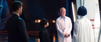 Movie still from “Barb and Star Go to Vista Del Mar” (2021), directed by Josh Greenbaum – A man in a lab coat stands next to a boy in a suit; Medium shot, Over the shoulder angle