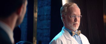 Movie still from “Barb and Star Go to Vista Del Mar” (2021), directed by Josh Greenbaum – An older man wearing a white lab coat and a bow tie; Close Up shot, Low angle