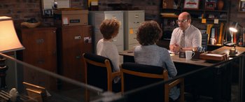 Movie still from “Barb and Star Go to Vista Del Mar” (2021), directed by Josh Greenbaum – Two women sitting at a table in front of filing cabinets; Wide shot, Over the shoulder angle