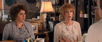 Movie still from “Barb and Star Go to Vista Del Mar” (2021), directed by Josh Greenbaum – Two women in a room with a lamp and a table in front of them; Close Up shot, Over the shoulder angle