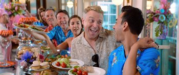 Movie still from “Barb and Star Go to Vista Del Mar” (2021), directed by Josh Greenbaum – A group of people sitting around a table with plates of food; Medium shot, Over the shoulder angle