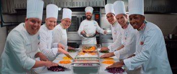 Movie still from “Barb and Star Go to Vista Del Mar” (2021), directed by Josh Greenbaum – A group of chefs preparing food in a commercial kitchen; Medium shot, High angle