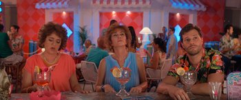 Movie still from “Barb and Star Go to Vista Del Mar” (2021), directed by Josh Greenbaum – A woman sitting at a table with a drink in a glass; Medium shot, Low angle