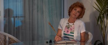 Movie still from “Barb and Star Go to Vista Del Mar” (2021), directed by Josh Greenbaum – An older woman sitting at a table with a feather pen; Medium shot, Over the shoulder angle