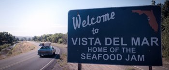 Movie still from “Barb and Star Go to Vista Del Mar” (2021), directed by Josh Greenbaum – A road sign that reads " welcome to vista del mar home of seafood "; Extreme Close Up shot, Low angle