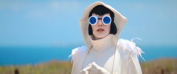Movie still from “Barb and Star Go to Vista Del Mar” (2021), directed by Josh Greenbaum – A woman in a white coat and blue sunglasses; Medium shot, Low angle