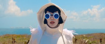 Movie still from “Barb and Star Go to Vista Del Mar” (2021), directed by Josh Greenbaum – A woman wearing a white coat and blue sunglasses; Close Up shot, Low angle