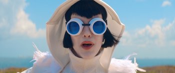 Movie still from “Barb and Star Go to Vista Del Mar” (2021), directed by Josh Greenbaum – A woman wearing a white coat and blue sunglasses; Close Up shot, Low angle