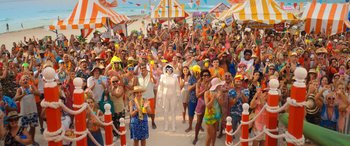 Movie still from “Barb and Star Go to Vista Del Mar” (2021), directed by Josh Greenbaum – A crowd of people standing around a man in a white suit; Wide shot, High angle