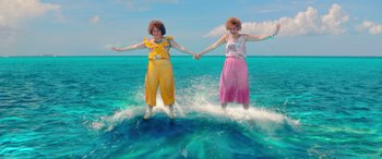 Movie still from “Barb and Star Go to Vista Del Mar” (2021), directed by Josh Greenbaum – A couple of women standing on a surfboard in the ocean; Wide shot, High angle