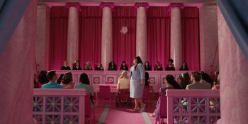 Movie still from “Barbie” (2023), directed by Greta Gerwig – A woman standing in front of a group of people in front of a pink wall; Extreme Wide shot, High angle