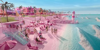 Movie still from “Barbie” (2023), directed by Greta Gerwig – A beach with people sitting on the sand and a lighthouse in the background; Extreme Wide shot, High angle