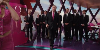 Movie still from “Barbie” (2023), directed by Greta Gerwig – A man in a suit and tie standing in front of a group of men; Wide shot, Low angle