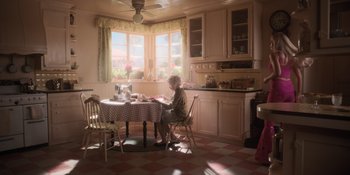 Movie still from “Barbie” (2023), directed by Greta Gerwig – An older woman sitting at a table in a kitchen; Wide shot, High angle