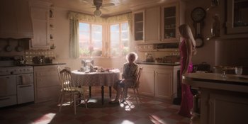 Movie still from “Barbie” (2023), directed by Greta Gerwig – An older woman sitting at a table in a kitchen; Wide shot, High angle