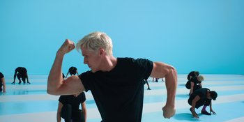 Movie still from “Barbie” (2023), directed by Greta Gerwig – A man with blond hair is stretching his arms; Medium shot, Low angle