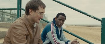 Movie still from “Empire of Light” (2022), directed by Sam Mendes – Two men sitting next to each other on the beach; Medium shot, Over the shoulder angle