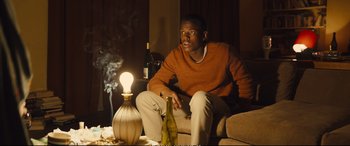 Movie still from “Empire of Light” (2022), directed by Sam Mendes – A man sitting on a couch next to a lamp and a bottle of wine; Medium shot, Low angle
