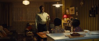 Movie still from “Empire of Light” (2022), directed by Sam Mendes – A man standing at a dining room table holding a fork; Medium shot, Over the shoulder angle