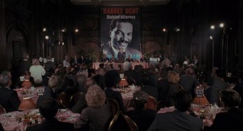 Movie still from “Batman” (1989), directed by Tim Burton – People sitting at tables in front of a poster of harvey dent; Wide shot, High angle