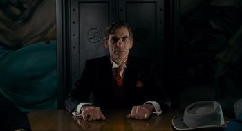 Movie still from “Batman” (1989), directed by Tim Burton – A man sitting at a table in front of a door; Medium shot, Low angle