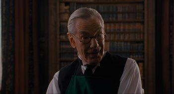 Movie still from “Batman” (1989), directed by Tim Burton – An older man wearing glasses and an apron in front of a book shelf; Close Up shot, Over the shoulder angle