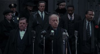 Movie still from “Batman” (1989), directed by Tim Burton – A group of people standing around a man holding microphones; Medium shot, Low angle