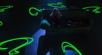 Movie still from “Batman Forever” (1995), directed by Joel Schumacher – A woman sitting on a couch in a room lit up with green neon lights; Wide shot, High angle