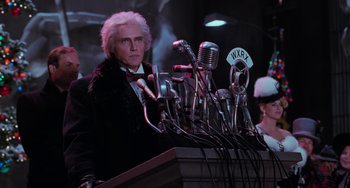 Movie still from “Batman Returns” (1992), directed by Tim Burton – A man in a suit and bow tie standing at a podium with many microphones; Medium shot, Low angle
