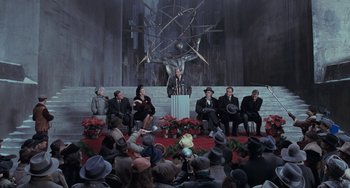 Movie still from “Batman Returns” (1992), directed by Tim Burton – A man is speaking at an event in front of a group of people; Wide shot, High angle