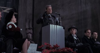 Movie still from “Batman Returns” (1992), directed by Tim Burton – A man is speaking at a podium while another man looks on; Medium shot, Low angle