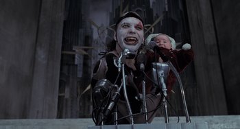Movie still from “Batman Returns” (1992), directed by Tim Burton – A man holding a baby in his arms while standing behind microphones and microphones; Medium shot, Low angle