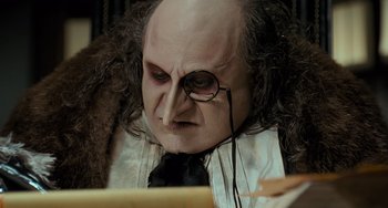 Movie still from “Batman Returns” (1992), directed by Tim Burton – A man with a bald head wearing a black tie and glasses; Close Up shot, Low angle