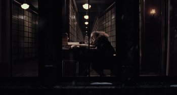 Movie still from “Batman Returns” (1992), directed by Tim Burton – A person sitting at a table in a dark room; Wide shot, High angle