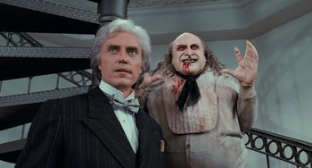 Movie still from “Batman Returns” (1992), directed by Tim Burton – A wax statue of a man and a man with blood on his face; Medium shot, Low angle