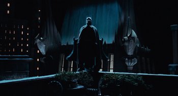 Movie still from “Batman Returns” (1992), directed by Tim Burton – A man standing in front of a stage with a bat on his back; Wide shot, Low angle