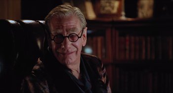 Movie still from “Batman & Robin” (1997), directed by Joel Schumacher – An older man wearing glasses sitting in a chair in a room; Close Up shot, Low angle