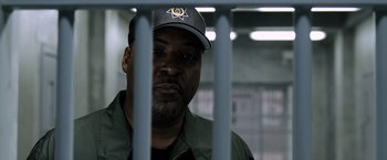 Movie still from “Batman v Superman: Dawn of Justice” (2016), directed by Zack Snyder – A man with a hat is behind bars in a jail cell; Close Up shot, Over the shoulder angle