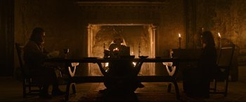 Movie still from “The Last Duel” (2021), directed by Ridley Scott – A person sitting at a table in front of a fireplace; Wide shot, Low angle