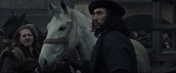 Movie still from “The Last Duel” (2021), directed by Ridley Scott – A man with long black hair standing next to a white horse; Close Up shot, Over the shoulder angle
