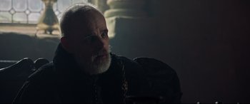 Movie still from “The Last Duel” (2021), directed by Ridley Scott – An old man with a beard and a mustache is sitting in a dark room; Close Up shot, Over the shoulder angle