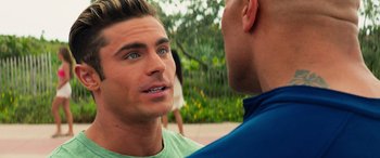 Movie still from “Baywatch” (2017), directed by Seth Gordon – Two men talking to each other in front of trees and flowers; Close Up shot, Over the shoulder angle