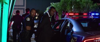 Movie still from “Baywatch” (2017), directed by Seth Gordon – A man in a suit and tie standing next to a police car; Medium shot, Over the shoulder angle