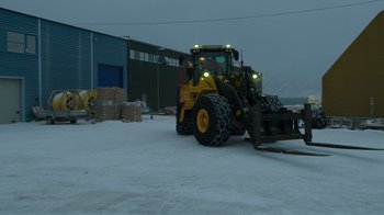 Movie still from “Bear Witness” (2022), directed by Jeff Wilson – A large yellow and black wheel loader is parked in the snow near a building; Extreme Wide shot, Over the shoulder angle