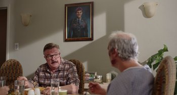 Movie still from “Beau Is Afraid” (2023), directed by Ari Aster – Two older men sitting at a table with a portrait of a soldier hanging on the wall behind them; Medium shot, Over the shoulder angle
