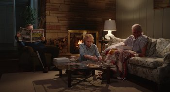 Movie still from “Beau Is Afraid” (2023), directed by Ari Aster – An older woman sitting at a coffee table in front of a fireplace; Wide shot, High angle